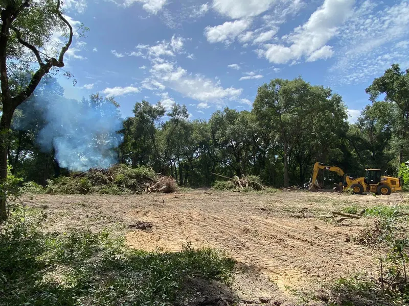 Cleared land with a controlled burn pile smoking at the forest edge and a CAT loader working in the background